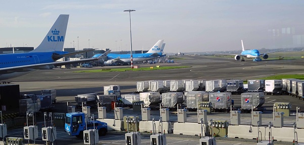 View Of Advertisement Sign, Cargo Containers On The Ground, Ground Crew With Pushback Tractor Towing KLM Airlines Passenger Airplane To Runway For Departure At Loading, Unloading Gate Of Amsterdam Schiphol International Airport In The Netherlands Europe