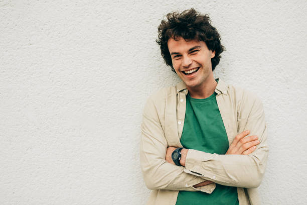 istock Handsome young man posing for advertisement, smiling and looking at the camera, standing at building concrete wall outdoors. Portrait of happy smart student male with curly hair with crossed arms. 1194460693