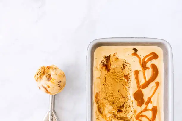 Homemade caramel ice cream with caramel sauce in a container and ice cream scoop on white marble background with copy space. Top view.