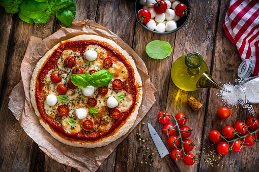 Italian food: delicious homemade pizza with mozzarella and cherry tomatoes shot from above on rustic wooden table. Some ingredients for preparing pizza like mozzarella cheese, basil, fresh tomatoes, olive oil, salt and pepper are all around the pizza. Predominant colors are red, white and brown. High resolution 42Mp studio digital capture taken with SONY A7rII and Zeiss Batis 40mm F2.0 CF