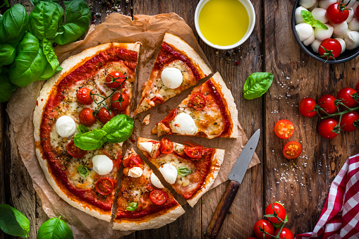 Italian food: delicious homemade pizza with mozzarella and cherry tomatoes shot from above on rustic wooden table. Some ingredients for preparing pizza like mozzarella cheese, basil, fresh tomatoes, olive oil, salt and pepper are all around the pizza. predominant colors are red, white and brown. High resolution 42Mp studio digital capture taken with SONY A7rII and Zeiss Batis 40mm F2.0 CF