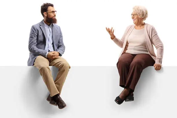 Senior lady sitting on a blank panel and talking to a younger bearded man isolated on white background