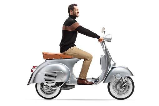 Full length profile shot of an elegant trendy man riding a silver vintage scooter isolated on white background
