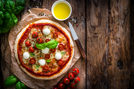 Italian food: delicious homemade pizza with mozzarella and cherry tomatoes shot from above on rustic wooden table. Some ingredients for preparing pizza like mozzarella cheese, basil, fresh tomatoes, olive oil, salt and pepper are all around the pizza. The composition is at the left of an horizontal frame leaving useful copy space for text and/or logo at the right. Predominant colors are red, white and brown. High resolution 42Mp studio digital capture taken with SONY A7rII and Zeiss Batis 40mm F2.0 CF