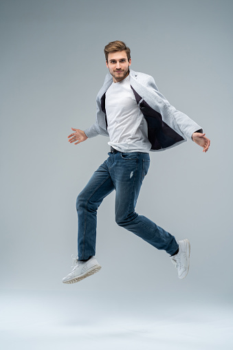 Full-length photo of funny man in casual t-shirt, blazer and jeans running or jumping in air isolated over gray background