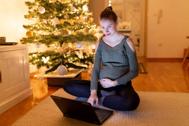 porträt der schönen schwangeren frau sitzt auf dem boden und arbeitet auf laptop mit weihnachtsbaum im hintergrund. - christmas shopping internet family stock-fotos und bilder