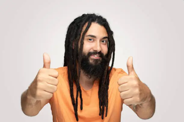 Confident handsome young man showing thumbs up over white background.He looking at camera with happy facial expression.Young handsome bearded man hippie with dreadlocks.Copyspace for ads.Studio shot, isolated background, real people ,horizontal composition.Young man wearing orange  tshirt and he has long and black hair.