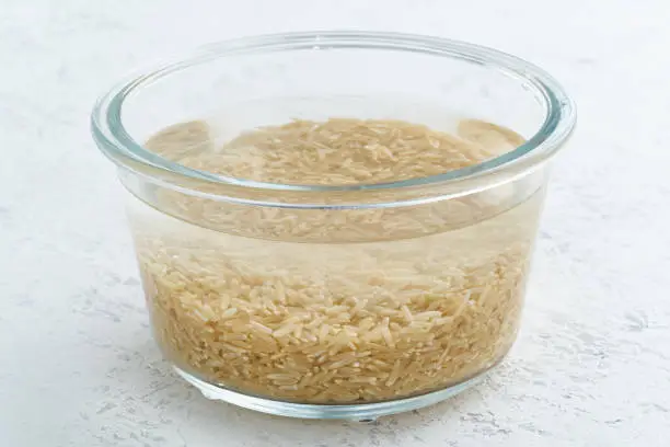 Soaking brown rice cereal in a water to ferment cereals and neutralize phytic acid. Large glass bowl with grains flooded with water. Side view, close up, white background