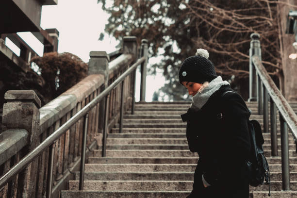 Woman wearing fur hat, warm coat and gloves sitting on snow-covered stone stairs in winter park. Cozy mood, cold sunny day, seasonal fashion and relaxed outdoor lifestyle concept