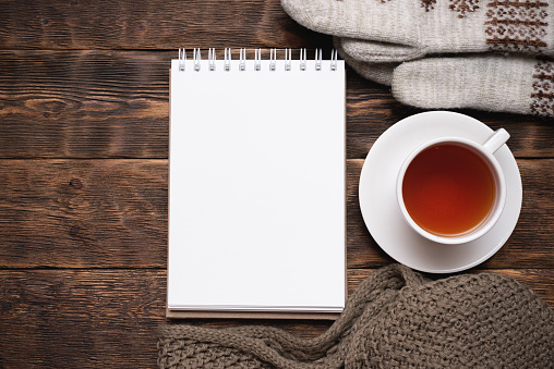 Blank notepad with copy space, warm clothes and cup of tea on brown wooden table flat lay background.
