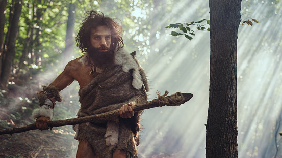 Portrait of Primeval Caveman Wearing Animal Skin and Fur Hunting with a Stone Tipped Spear in the Prehistoric Forest. Prehistoric Neanderthal Hunter Scavenging with Primitive Tools in the Jungle