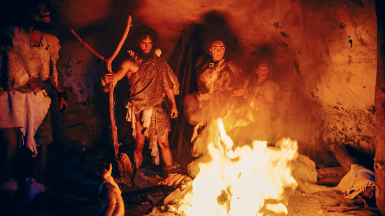 Tribu de cazadores-recolectores prehistóricos que llevan pieles de animales se paran alrededor de la hoguera fuera de la cueva por la noche. Retrato de Neanderthal / Homo Sapiens Familia Haciendo Ritual de Religión Pagana Cerca del Fuego photo