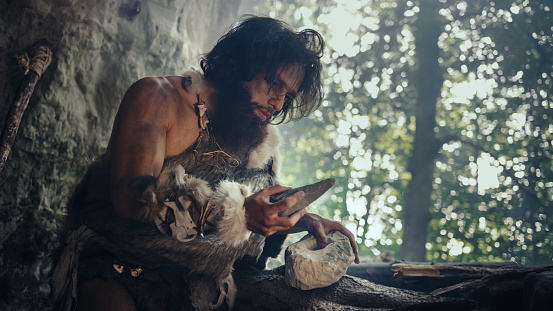 Primeval Caveman Wearing Animal Skin sostiene piedra afilada y hace la primera herramienta primitiva para cazar presas de animales, o para manejar pieles. Neandertal usando Handax. Amanecer de la civilización humana photo