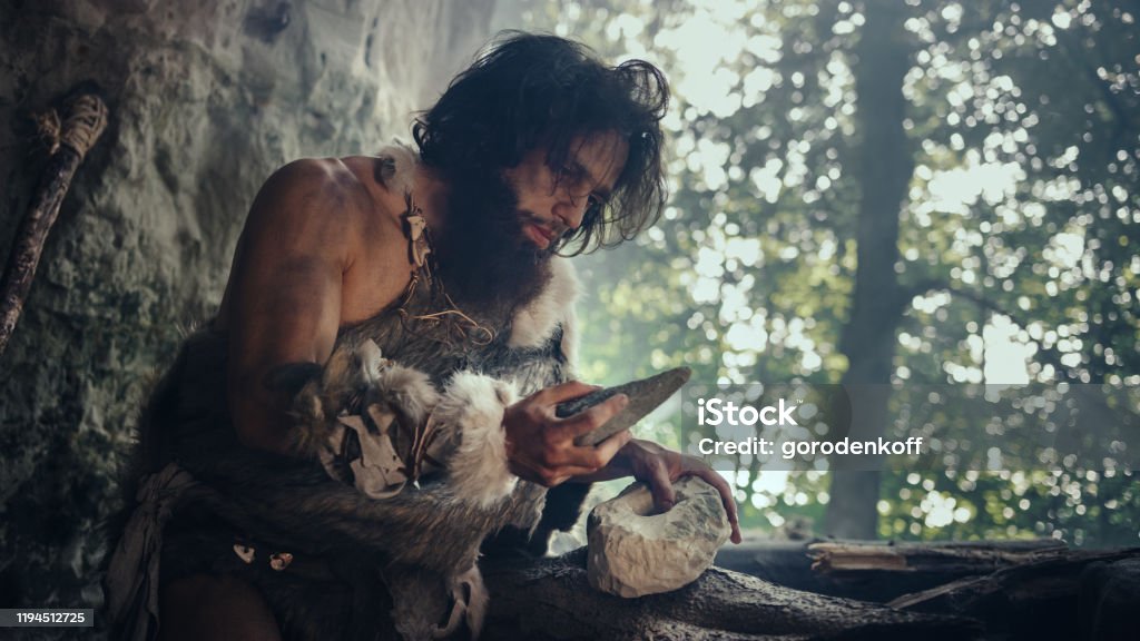 Primeval Caveman Wearing Animal Skin Holds Sharp Stone and Makes First Primitive Tool for Hunting Animal Prey, or to Handle Hides. Neanderthal Using Handax. Dawn of Human Civilization Neanderthal Stock Photo