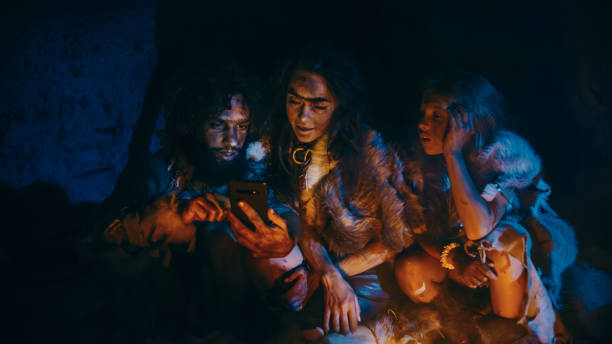 Tribe of Prehistoric, Primitive Hunter-Gatherers Wearing Animal Skins Use Smartphone in a Cave at Night. Neanderthal / Homo Sapiens Family Browsing Internet on Mobile Phone, Cooking Food over Fire tribu de prehistóricos, primitivocazadores cazadores que usan pieles de animales utilizan smartphone en una cueva en la noche. neanderthal / homo sapiens familia navegación por internet en el teléfono móvil, cocinar alimentos a través del fuego