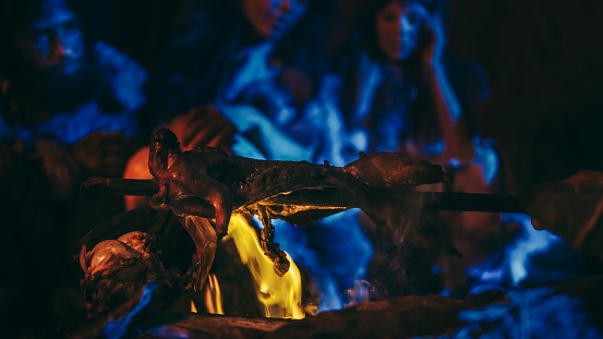 Neanderthal or Homo Sapiens Family Cooking Animal Meat over Bonfire and then Eating it. Tribe of Prehistoric Hunter-Gatherers Wearing Animal Skins Grilling and Eating Meat in Cave at Night