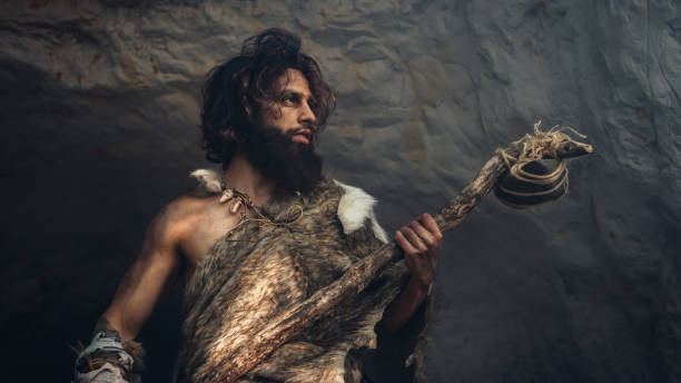 Primeval Caveman Wearing Animal Skin Holds Stone Hammer Stands Near Cave and Looks Around Prehistoric Landscape, Ready to Hunt Animal Prey. Neanderthal Going Hunting into Jungle. Low Angle Shot el cavernícola primitivo que lleva la piel de un animal sostiene un martillo de piedra cerca de la cueva y mira alrededor del paisaje prehistórico, listo para cazar presas de animales. neanderthal going hunting into jungle. disparo de ángulo bajo