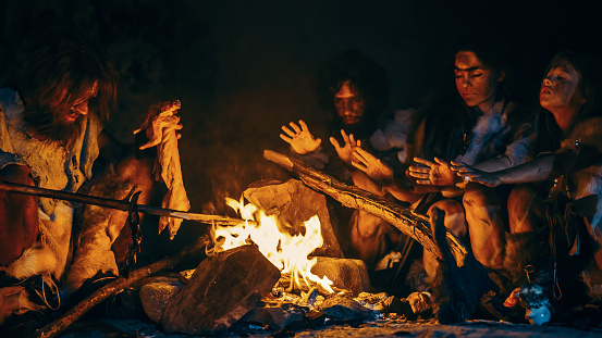 Neanderthal u Homo Sapiens Family Cooking Animal Meat over Bonfire y luego Comerla. Tribu de cazadores-recolectores prehistóricos que usan pieles de animales comiendo en una cueva de miedo oscuro por la noche photo