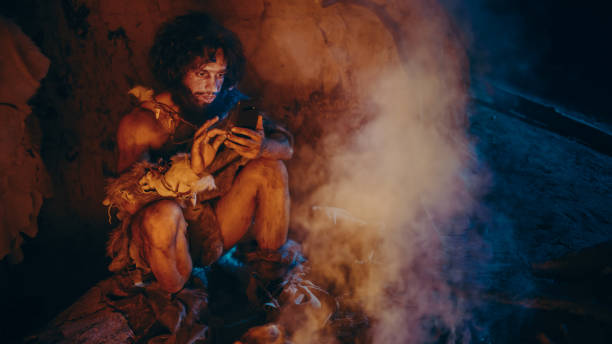 stamm der prähistorischen, primitive hunter gatherer trägt tierhaut verwendet smartphone in einer höhle in der nacht. neandertaler / homo sapiens männlich browsing internet auf handy, schaut videos - historical person stock-fotos und bilder