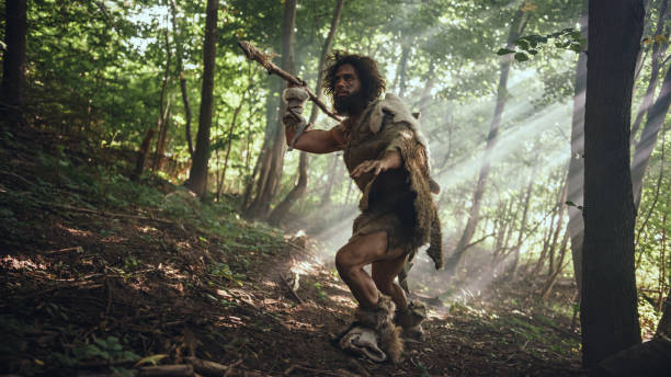 il cavernicolo primordiale che indossa la pelle animale tiene la lancia con punta di pietra guarda intorno, esplora la foresta preistorica in una caccia alla preda animale. neanderthal va a caccia nella giungla - neanderthal foto e immagini stock