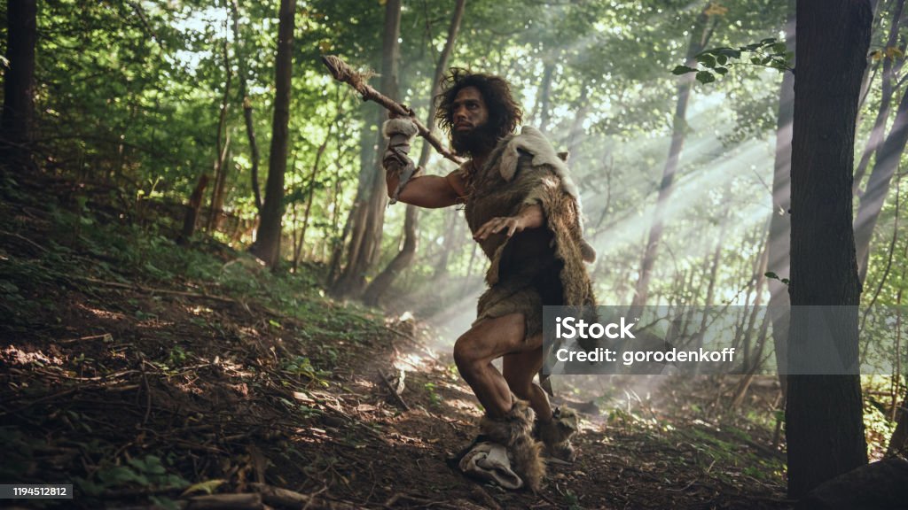 Primeval Caveman Wearing Animal Skin Holds Stone Tipped Spear Looks Around, Explores Prehistoric Forest in a Hunt for Animal Prey. Neanderthal Going Hunting in the Jungle Caveman Stock Photo