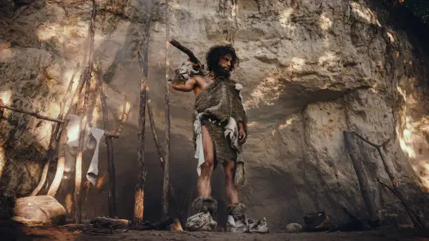 Primeval Caveman Wearing Animal Skin Holds Stone Tipped Hammer Looks Around Prehistoric Forest, Ready to Hunt Animal Prey. Neanderthal Going Hunting into the Jungle.