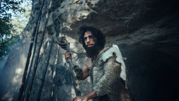 Primeval Caveman Wearing Animal Skin Holds Stone Hammer Stands Near Cave and Looks Around Prehistoric Landscape, Ready to Hunt Animal Prey. Neanderthal Going Hunting into Jungle. Low Angle Shot el cavernícola primitivo que lleva la piel de un animal sostiene un martillo de piedra cerca de la cueva y mira alrededor del paisaje prehistórico, listo para cazar presas de animales. neanderthal going hunting into jungle. disparo de ángulo bajo