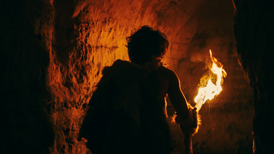 Primeval Caveman Wearing Animal Skin Exploring Cave At Night Holding Torch with Fire Looking at Drawings on the Walls at Night. Neanderthal Searching Safe Place to Spend the Night. Back View