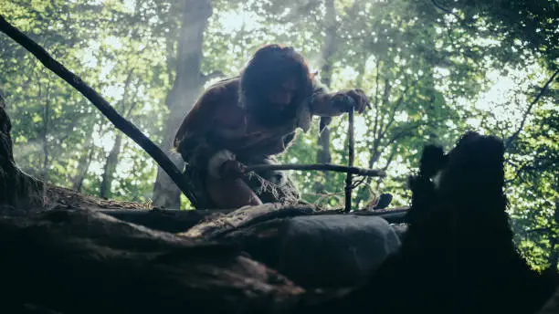 Silhouette of Primeval Caveman Wearing Animal Skin Trying to make a Fire with Bow Drill Method. Neanderthal Kindle First Man-Made fire in the Human Civilization History. Low Angle Shot