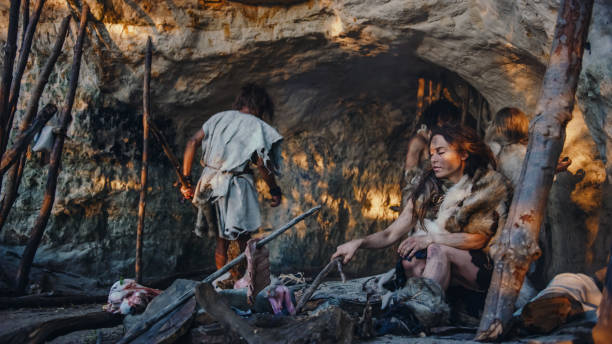 tribu de cazadores-recolectores que llevan piel de animal viven en una cueva. líder trae presas animales de la caza, cocinas femeninas de comida en la hoguera, niña dibujo en wals creando arte. neanderthal homo sapiens family - ancient fotografías e imágenes de stock