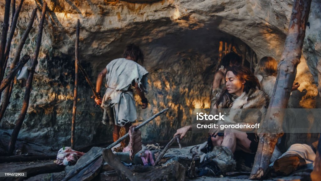Tribe of Hunter-Gatherers Wearing Animal Skin Live in a Cave. Leader Brings Animal Prey from Hunting, Female Cooks Food on Bonfire, Girl Drawing on Wals Creating Art. Neanderthal Homo Sapiens Family Prehistoric Era Stock Photo
