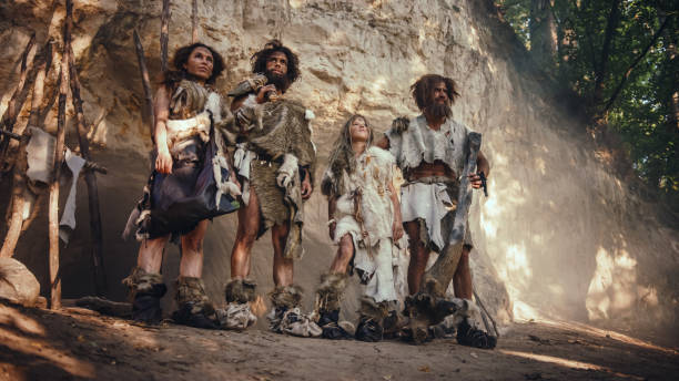 tribe of four hunter-gatherers wearing animal skin holding stone tipped tools, pose at the entrance of their cave. portrait of two grown male and two female neanderthals and their way of living - neanderthal imagens e fotografias de stock