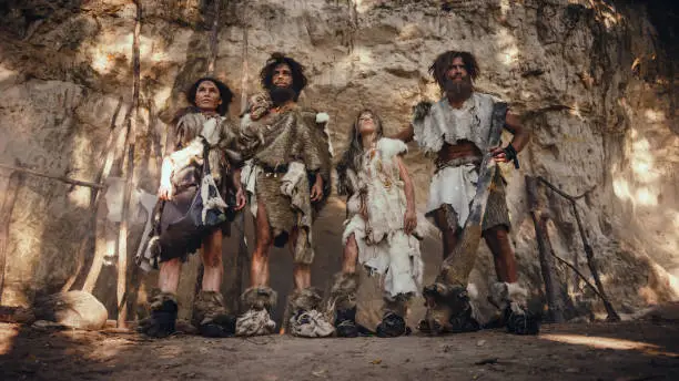 Photo of Tribe of Four Hunter-Gatherers Wearing Animal Skin Holding Stone Tipped Tools, Pose at the Entrance of their Cave. Portrait of Two Grown Male and Two Female Neanderthals and their Way of Living