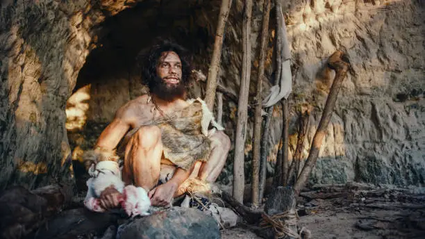 Photo of Primeval Caveman Wearing Animal Skin Holds Bone and Hits Rock with It. Neanderthal Fooling Around near the Cave Entrance, Maybe Creating first Primitive Tools or Weapons by Accident.