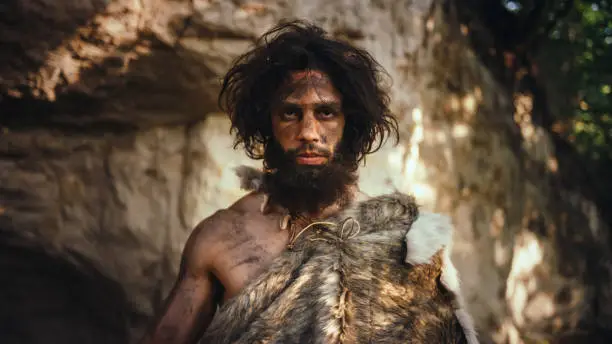Portrait of Primeval Caveman Wearing Animal Skin Holding Stone Tipped Hammer. Prehistoric Neanderthal Hunter Posing with Primitive Hunting in the Jungle. Looking at Camera