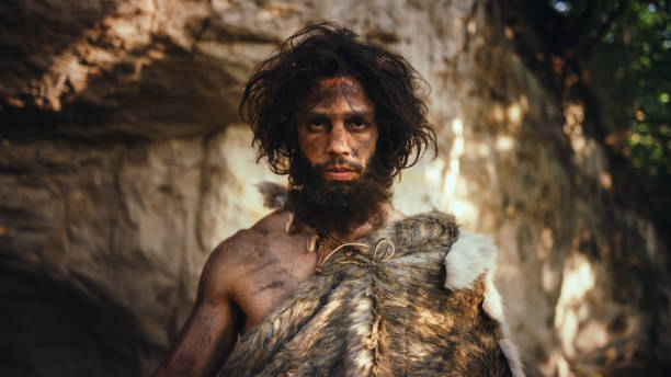 portrait of primeval caveman wearing animal skin holding stone tipped hammer. prehistoric neanderthal hunter posing with primitive hunting in the jungle. looking at camera - neanderthal imagens e fotografias de stock