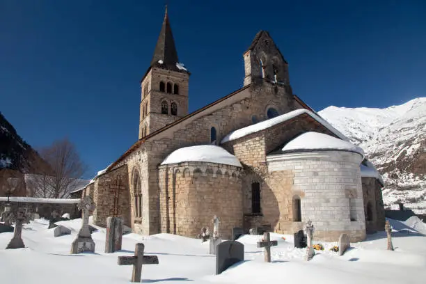 Horizontal exterior view of the Romanesque Santa María church with its apse and graveyard covered by snow, Artíes, Vall d’Aran, Lleida, Catalonia