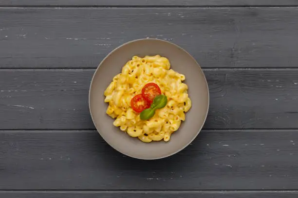 A bowl of delicious Macaroni and cheese, topped with cherry tomatoes and basil, on a distressed dark wooden background.