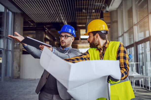Upset architect arguing with construction worker and showing him his mistake. Worker holding blueprints and defending himself. Building in construction process interior.