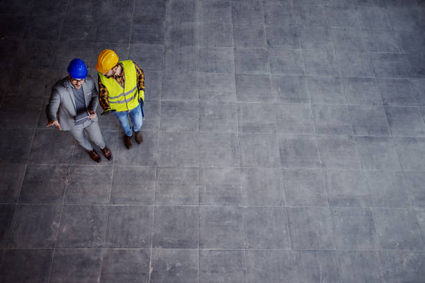 istock Top view of supervisor with helmet on head in suit standing with construction worker, holding tablet and explaining him ideas about project. 1194554380