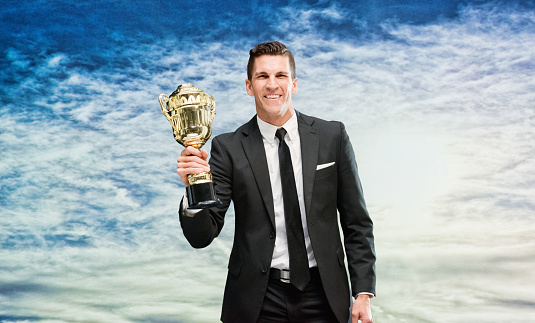 One man only / one person / waist up / front view / looking at camera of handsome people caucasian male / young men businessman / business person / manager standing in front of nature / scenics - nature who is outdoors wearing businesswear / a suit who is smiling / happy / cheerful / successful and winning / showing award who is in first place and holding trophy - award / gold / medal / sky
