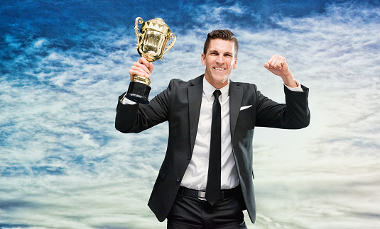 One man only / one person / waist up / front view / looking at camera of male / young men businessman / business person standing in front of nature / scenics - nature who is outdoors wearing businesswear who is smiling / happy / cheerful / excited / successful and cheering / winning / showing fist / award who is and doing fist pump / in first place and holding trophy - award / gold / medal / sky