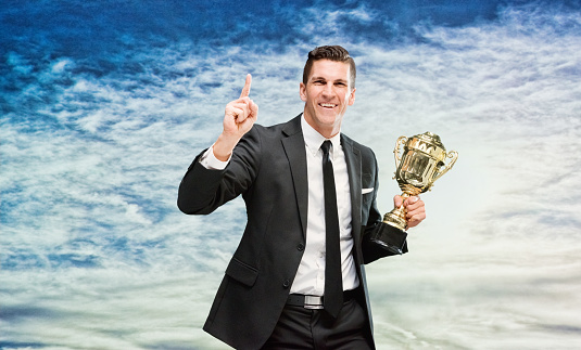 One man only / one person / waist up / front view of handsome people caucasian male / young men businessman / business person standing in front of nature / scenics - nature who is outdoors wearing businesswear / a suit who is smiling / happy / cheerful / successful and winning / showing award who is pointing / in first place and holding trophy - award / gold / medal / sky