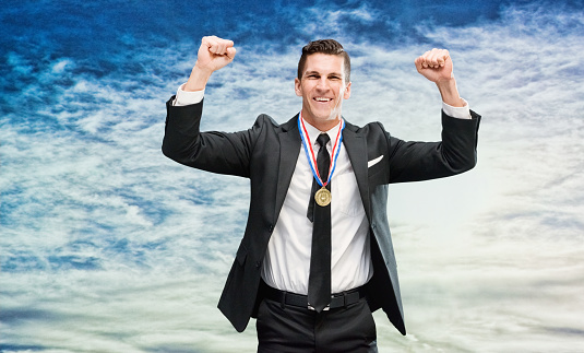 One man only / one person / waist up / front view of male / young men businessman / business person standing in front of nature / scenics - nature who is outdoors wearing businesswear who is smiling / happy / cheerful / excited / successful and cheering / showing fist / award who is and doing fist pump / in first place and holding medallion / gold medal / medal / gold / sky