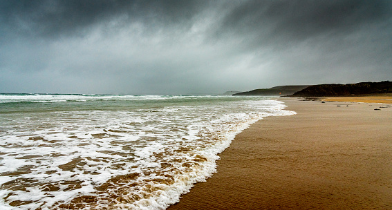 The white foam of the waves of the ocean comes to die on the sand under a dark sky laden with rain.