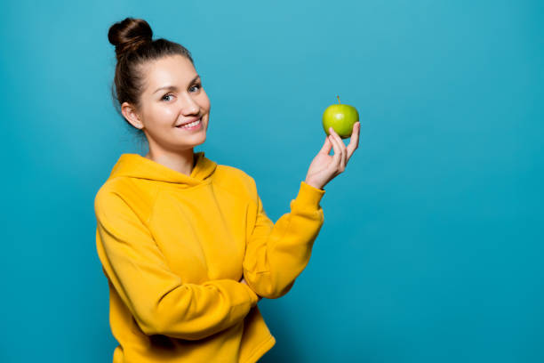 chica sonriente en una sudadera sostiene una manzana en la mano - apple women green eating fotografías e imágenes de stock