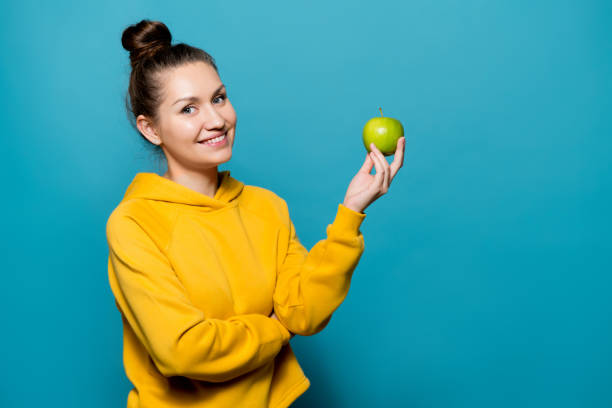 улыбающаяся девушка в толстовке держит яблоко в руке - apple women green eating стоковые фото и изображения