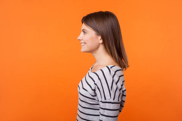 긴 소매 줄무늬 셔츠에 갈색 머리와 사랑스러운 행복한 젊은 여성의 사이드 뷰 초상화. 오렌지 배경에 고립 된 실내 스튜디오 샷 - side view 이미지 뉴스 사진 이미지