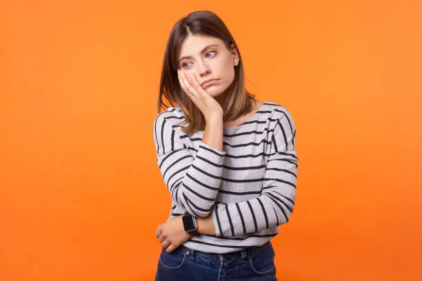 Portrait of bored young woman with brown hair in long sleeve shirt standing with face in hand, thinking sadly, looking depressed and disinterested. indoor studio shot isolated on orange background