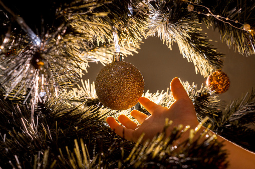 Kids hand touches gold christmas ball hanging on a Christmas tree. Beautiful back light.